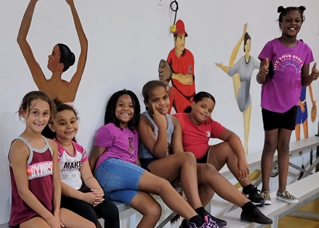 A group of young girls sit on bleachers inside a gymnasium.