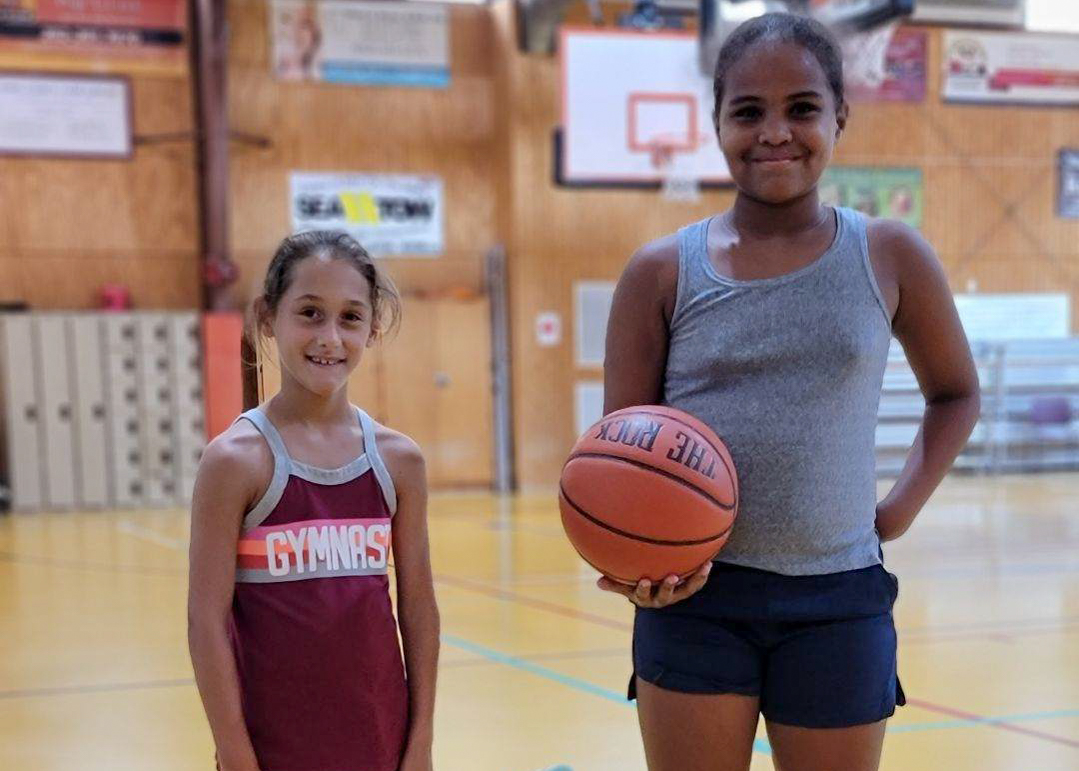 Two young girls smile at the camera. One holds a basketball.