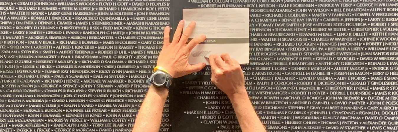 Image of an individual taking a name rubbing using paper and pencil against The Wall That Heals.