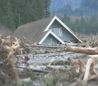 Image of a destroyed home in the mountains that is surrounded by flood water and debris