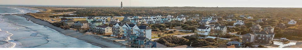 Aerial image of houses along the ocean in Buxton, featuring the Hatteras Lighthouse in the background.